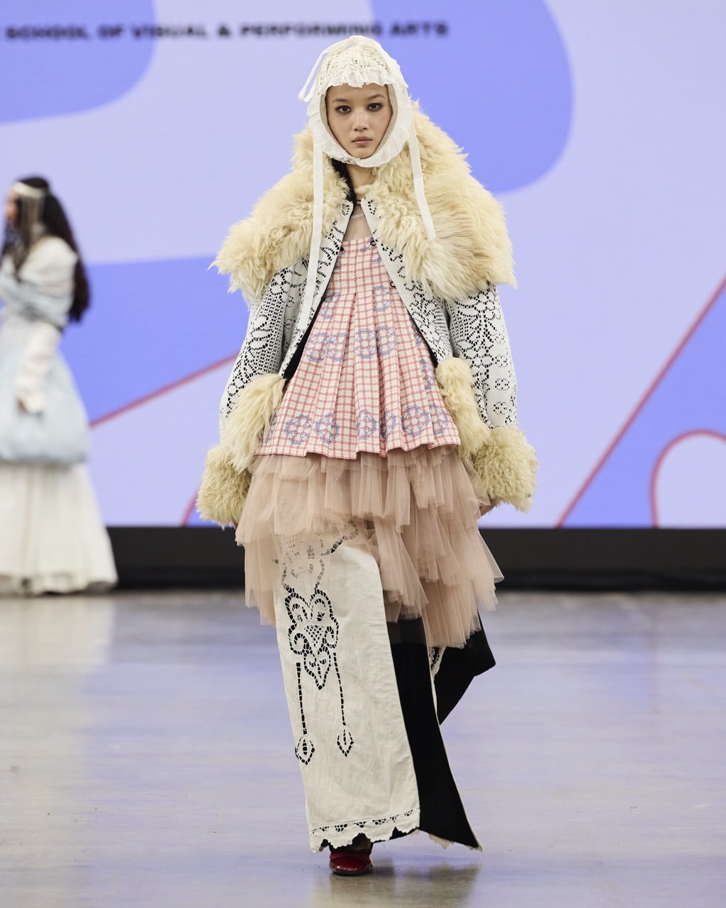 A model walks the runway in a layered outfit with a fur-trimmed jacket, a pink-chequered top, and patterned white and beige skirts during Graduate Fashion Week 2024.