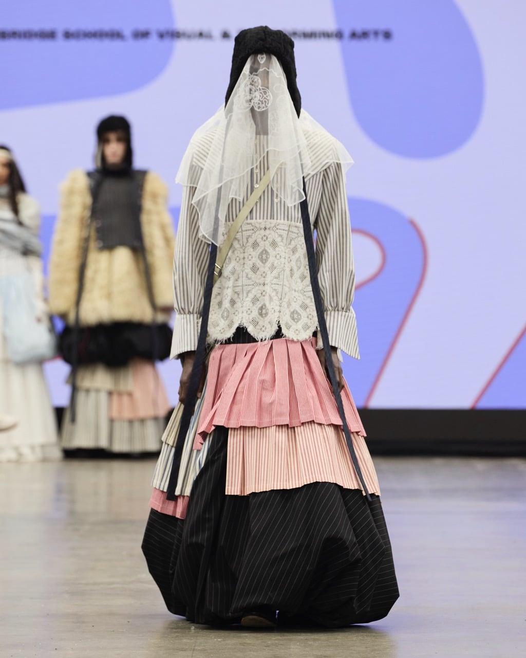 Model wearing a multi-layered dress with black, white, pink, and striped patterns, and a headpiece with white lace face covering, on a catwalk.