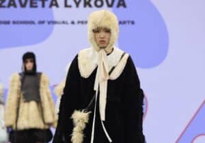 A model walking on a runway wearing a black-and-white outfit with a fur hat and a large bow, against a background with other models and a screen displaying text.