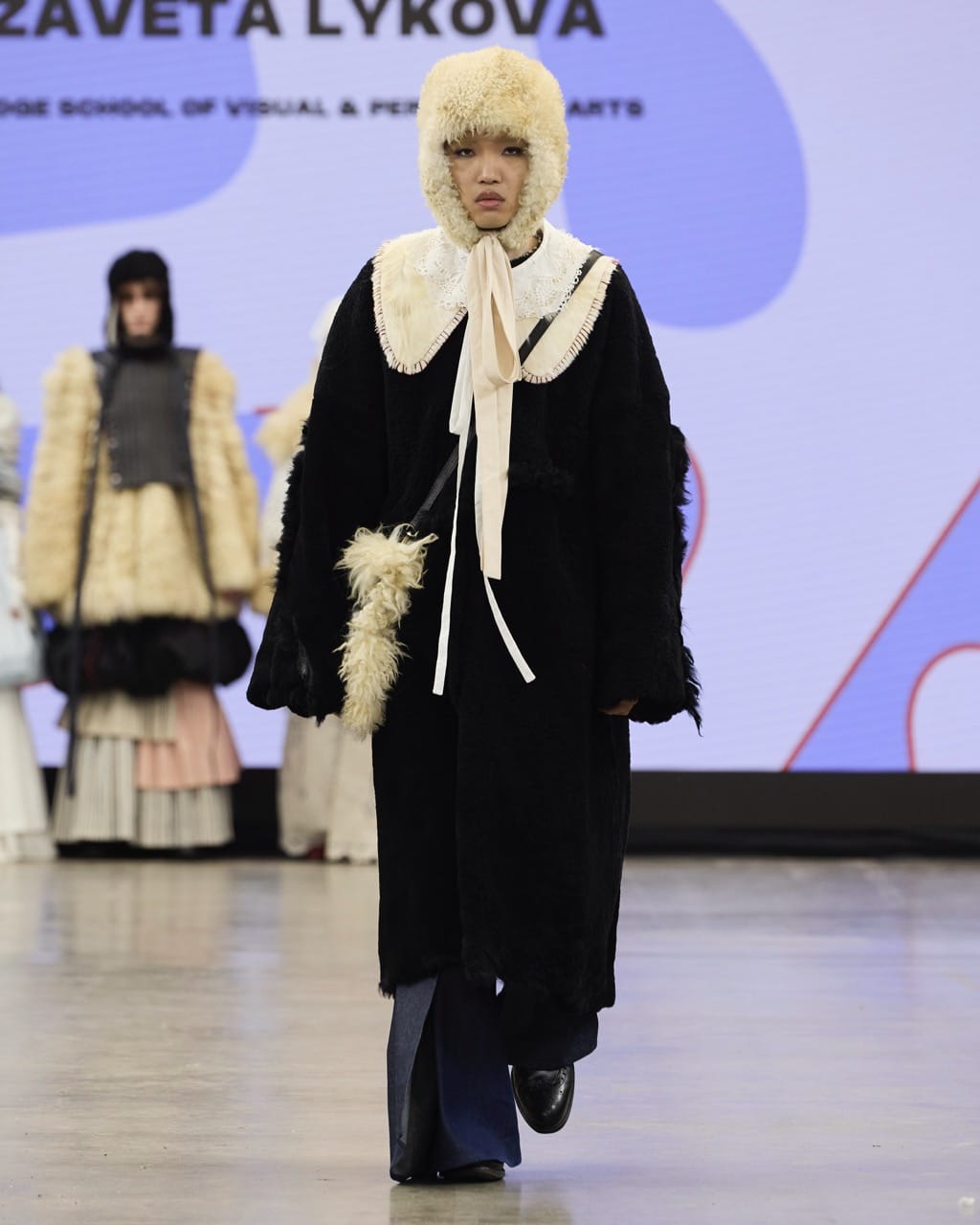 A model walking on a runway wearing a black-and-white outfit with a fur hat and a large bow, against a background with other models and a screen displaying text.