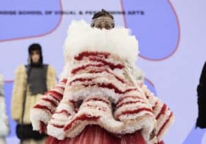 A model walks down the runway in an avant-garde outfit featuring layers of red and white ruffled fabric, a lace skirt, and a headpiece, presented at Graduate Fashion Week 2024.