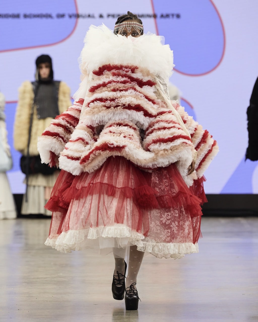 A model walks down the runway in an avant-garde outfit featuring layers of red and white ruffled fabric, a lace skirt, and a headpiece, presented at Graduate Fashion Week 2024.