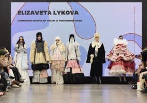 Seven models wearing layered, avant-garde fashion designs stand on a runway stage. A large screen behind them displays "Elizaveta Lykova, Cambridge School of Visual & Performing Arts" text.