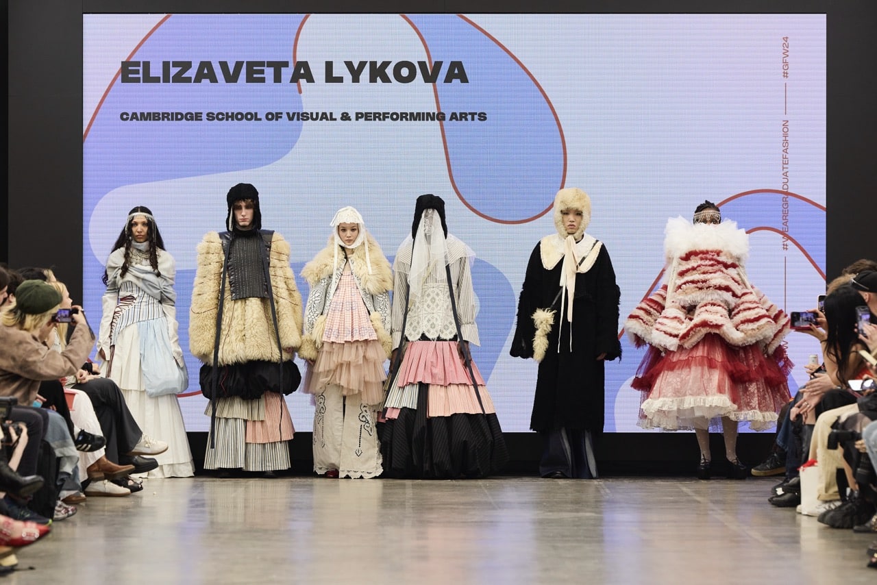 Seven models wearing layered, avant-garde fashion designs stand on a runway stage. A large screen behind them displays "Elizaveta Lykova, Cambridge School of Visual & Performing Arts" text.