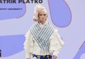 A model walks on a runway, wearing a multi-layered, avant-garde outfit with a mix of blue, red, and white patterns, and net-like adornments.