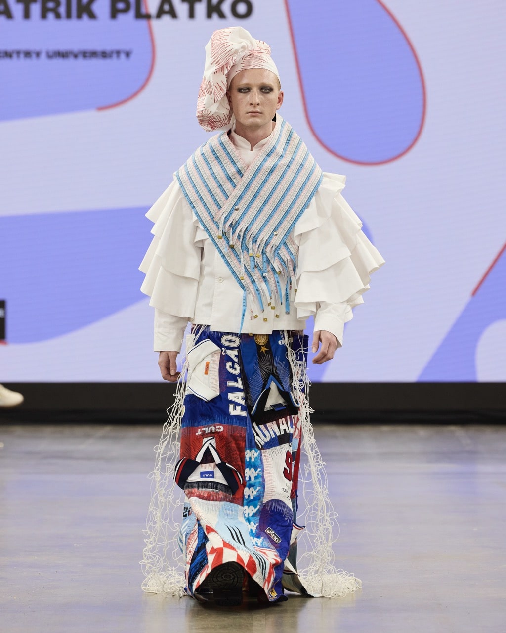 A model walks on a runway, wearing a multi-layered, avant-garde outfit with a mix of blue, red, and white patterns, and net-like adornments.