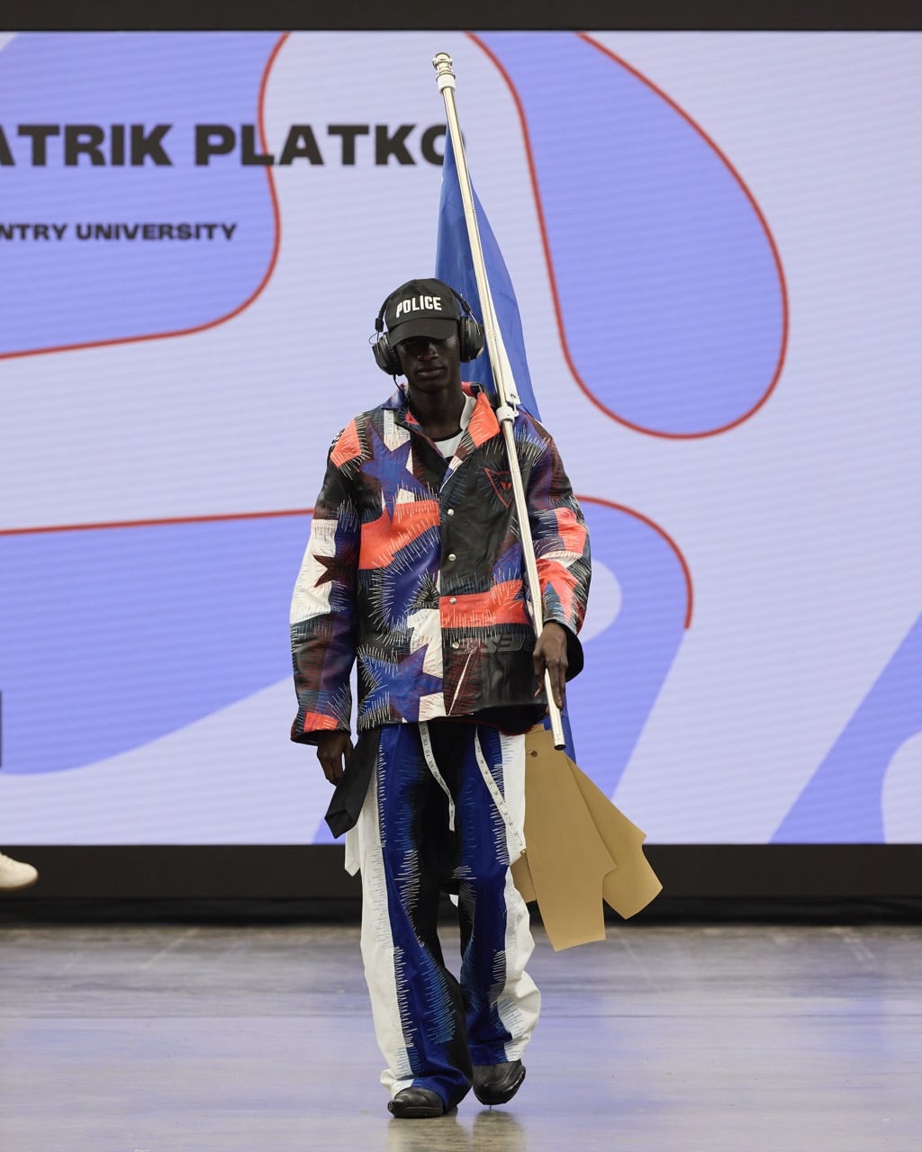 A person walking on a stage holding a flagpole. They are wearing a colourful, patterned outfit with a jacket and trousers, along with a cap that reads "POLICE" and headphones. A large screen in the background displays Graduate Fashion Week 2024.