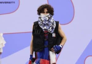 A model on a runway at Graduate Fashion Week 2024 wearing a black vest, a camo face covering, and a colourful skirt with "POLICE" text, set against a blue and white backdrop.