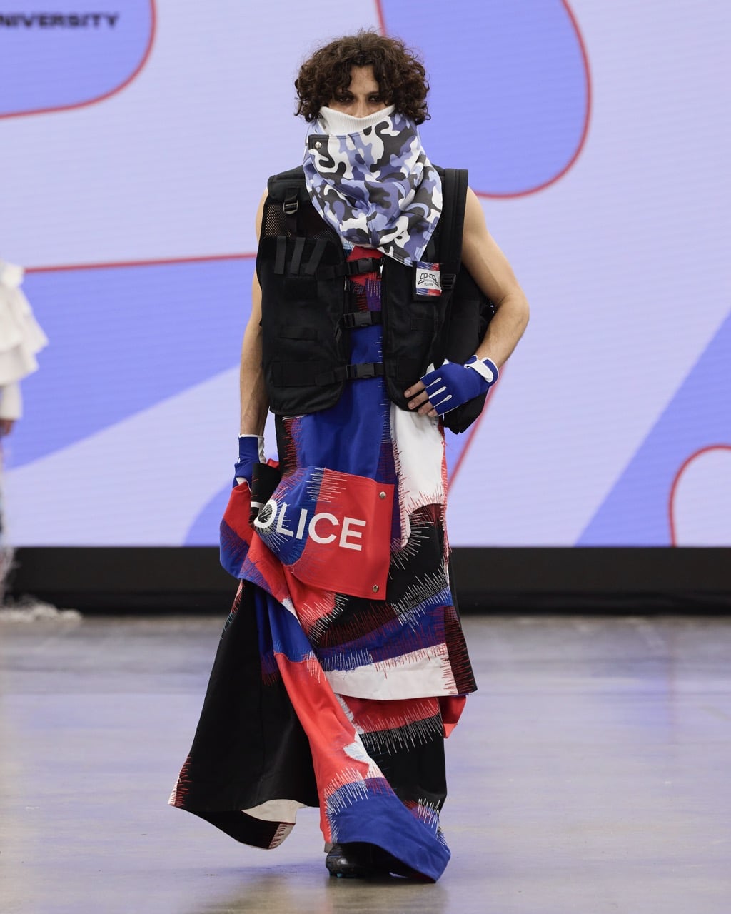 A model on a runway at Graduate Fashion Week 2024 wearing a black vest, a camo face covering, and a colourful skirt with "POLICE" text, set against a blue and white backdrop.