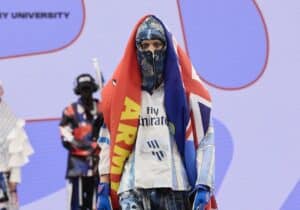 A model in vibrant, eclectic clothing including a headscarf, cape, and sports jersey walks down the runway at Graduate Fashion Week 2024. Other models and a backdrop with text are visible in the background.