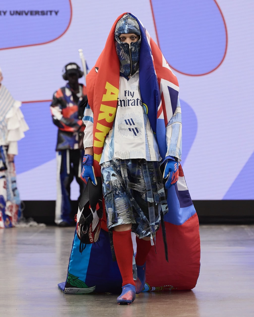 A model in vibrant, eclectic clothing including a headscarf, cape, and sports jersey walks down the runway at Graduate Fashion Week 2024. Other models and a backdrop with text are visible in the background.