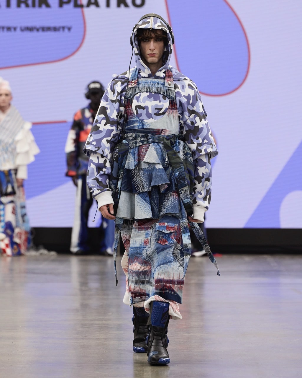 A model on a runway wearing a multi-layered outfit featuring a mix of camouflage and patchwork denim patterns.