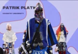 A model walks the runway wearing a colourful, multi-layered outfit and helmet during Patrik Platko's fashion show at Coventry University, part of Graduate Fashion Week 2024. A screen with Patrik Platko's name is displayed in the background.