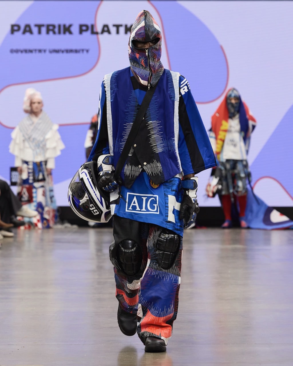 A model walks the runway wearing a colourful, multi-layered outfit and helmet during Patrik Platko's fashion show at Coventry University, part of Graduate Fashion Week 2024. A screen with Patrik Platko's name is displayed in the background.