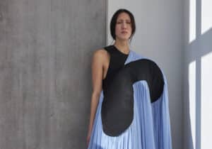 A person stands against a wall wearing a flowing blue dress with a unique black design, extending to the floor. They have long dark hair pulled back, and the scene is lit by natural window light.