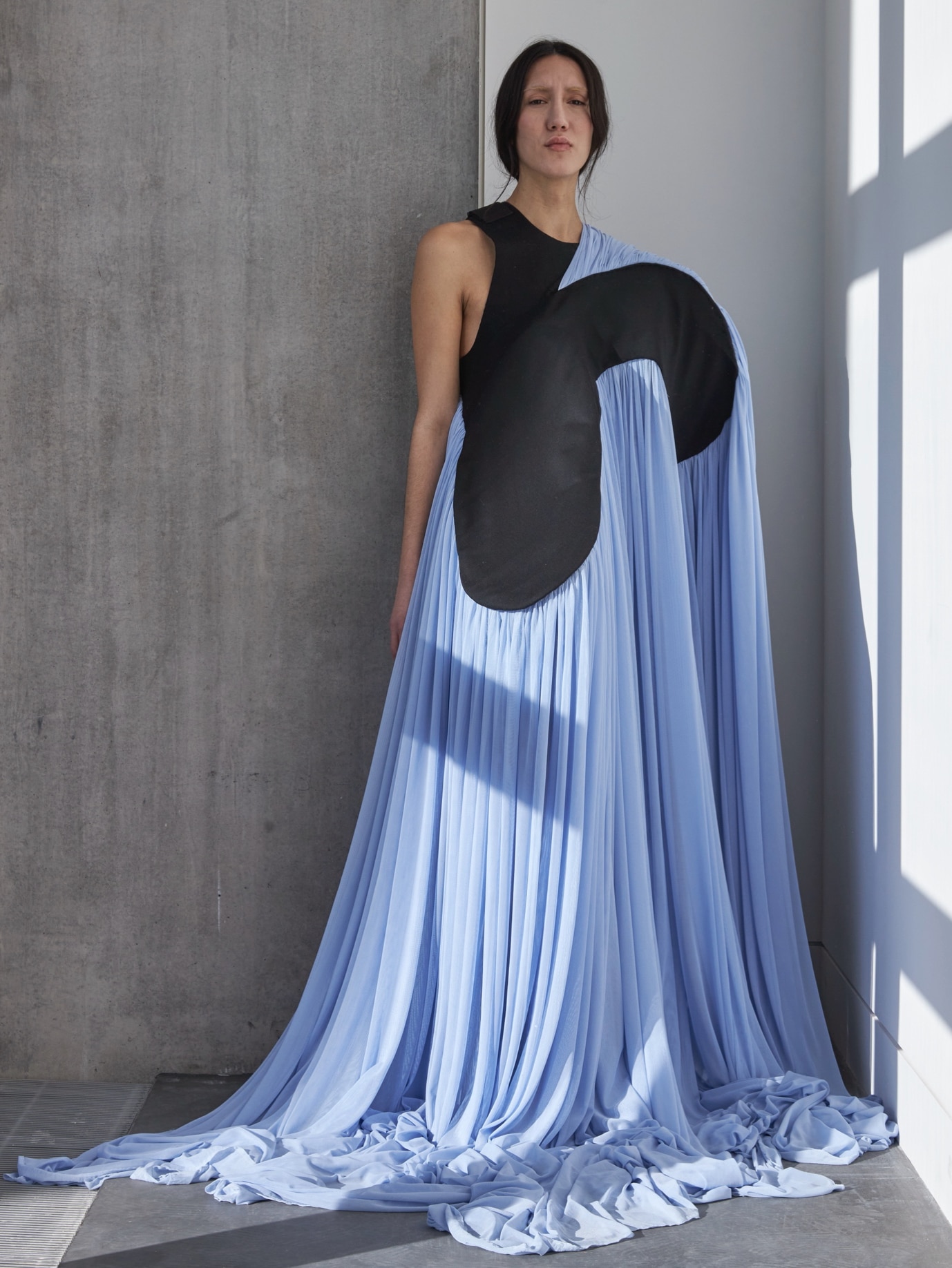 A person stands against a wall wearing a flowing blue dress with a unique black design, extending to the floor. They have long dark hair pulled back, and the scene is lit by natural window light.