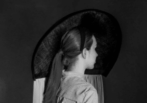 A black and white image of a person wearing a striped outfit, a large hat with a circular design, and a ponytail. The person is facing sideways against a dark background.