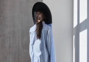 A person stands facing a wall, wearing a stripy shirt, black trousers, and a large black hat with long flowing blue fabric attached to the back.