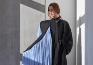 A person wearing a black coat with a long, flowing, light blue fabric draped from it stands against a concrete wall in a well-lit room.