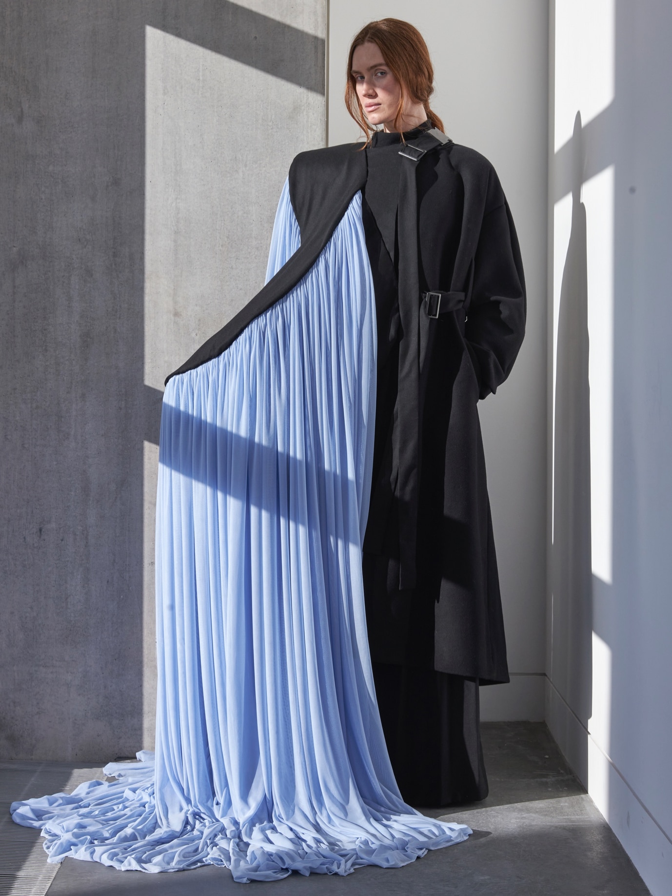 A person wearing a black coat with a long, flowing, light blue fabric draped from it stands against a concrete wall in a well-lit room.