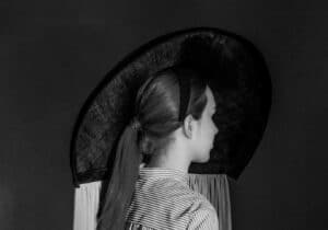 A person with long hair tied back is seen from behind wearing a large, wide-brimmed hat and a striped outfit with a circular cut-out on the back.