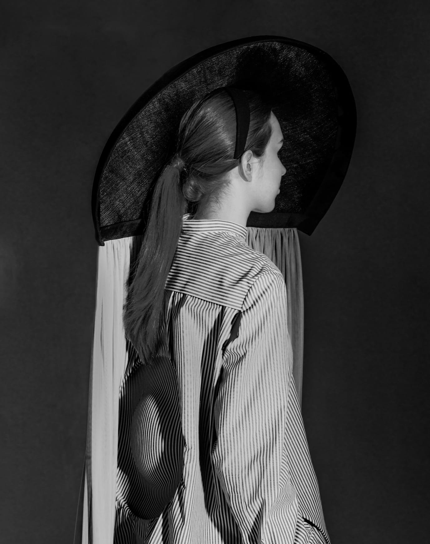 A person with long hair tied back is seen from behind wearing a large, wide-brimmed hat and a striped outfit with a circular cut-out on the back.