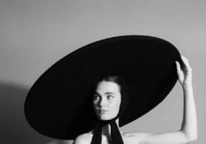 A woman in a strapless black and white dress with a voluminous skirt and a large round black hat poses against a plain background, holding the edge of the hat with one hand.