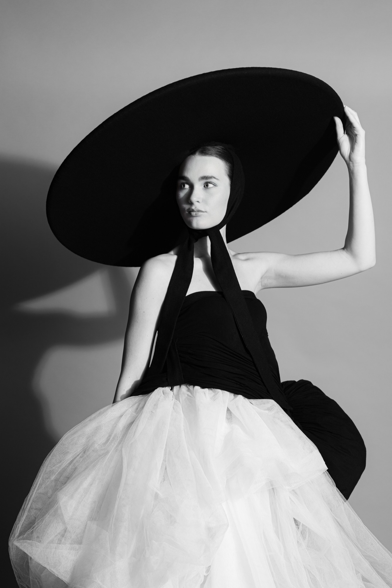 A woman in a strapless black and white dress with a voluminous skirt and a large round black hat poses against a plain background, holding the edge of the hat with one hand.