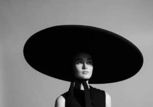 A monochrome image of a woman wearing a large, round hat and a long, sleeveless black dress, standing against a plain background.