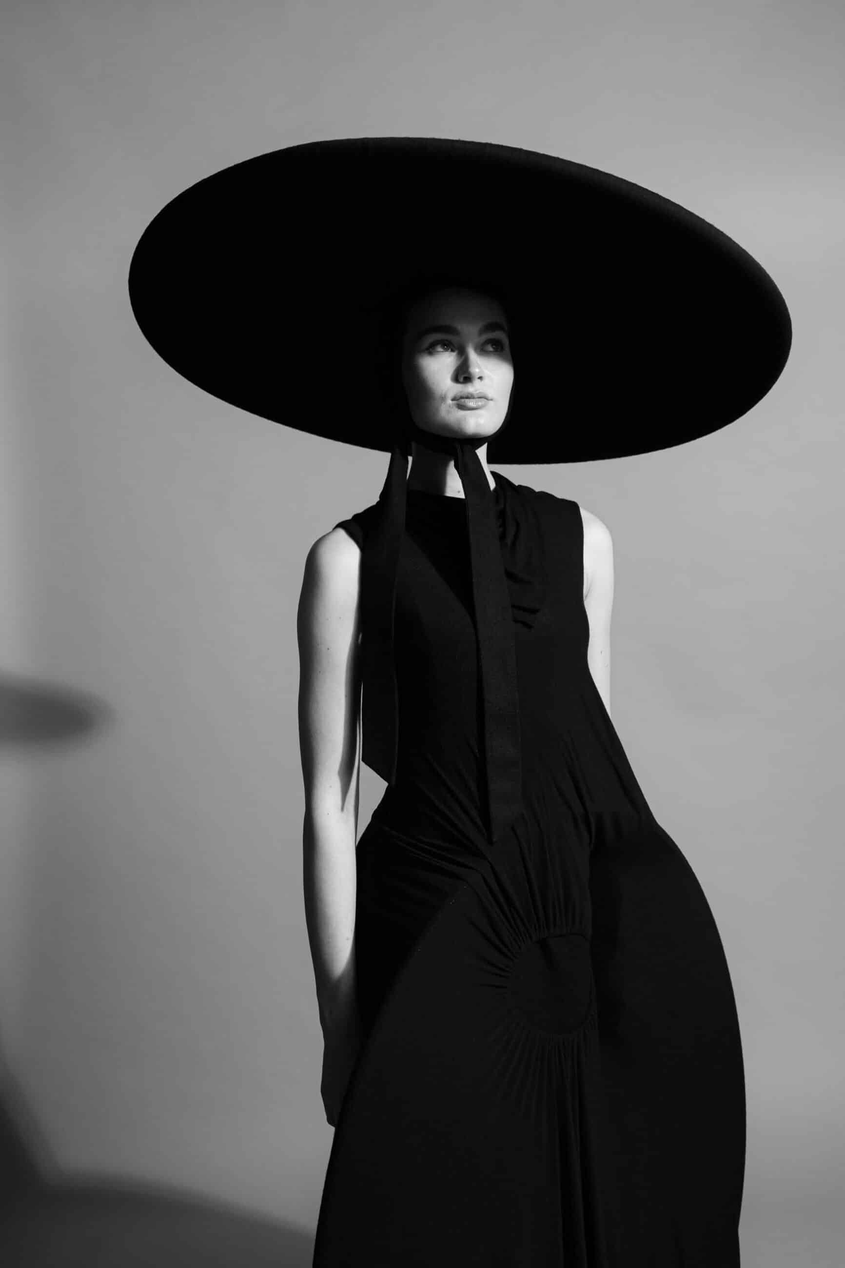 A monochrome image of a woman wearing a large, round hat and a long, sleeveless black dress, standing against a plain background.