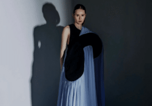 A person stands against a plain wall, wearing a dramatic gown with a black top and long, flowing blue fabric extending onto the floor.