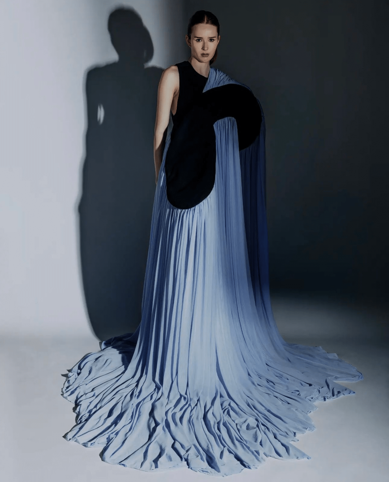 A person stands against a plain wall, wearing a dramatic gown with a black top and long, flowing blue fabric extending onto the floor.