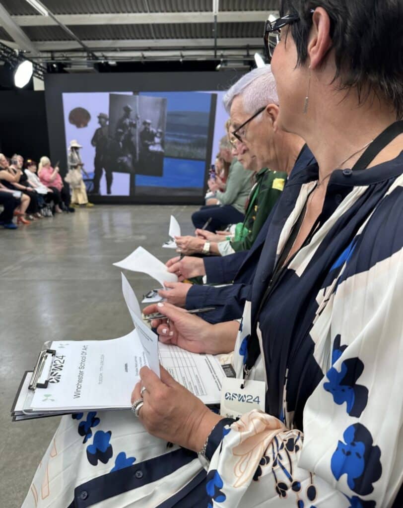 People seated in a row, holding clipboards and papers, at a fashion event with a runway.