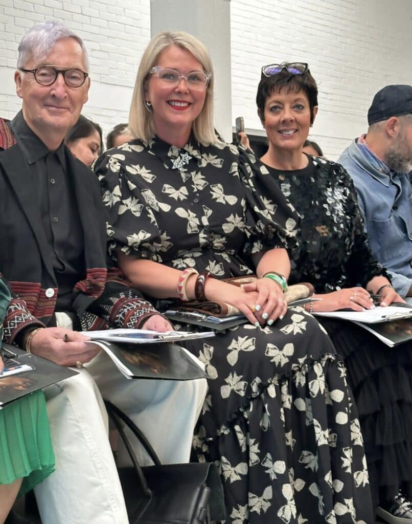 Three judges sit together at Graduate Fashion Week 2024, smiling. Two women in floral dresses are in the centre, with a man in a patterned jacket and white trousers on the left.