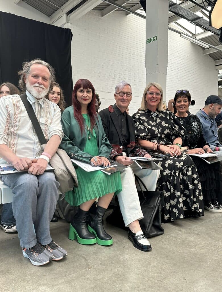 A group of five people seated side by side in a bright, open space, dressed in diverse stylish outfits, holding open programmes or magazines. Others are seated and standing in the background.