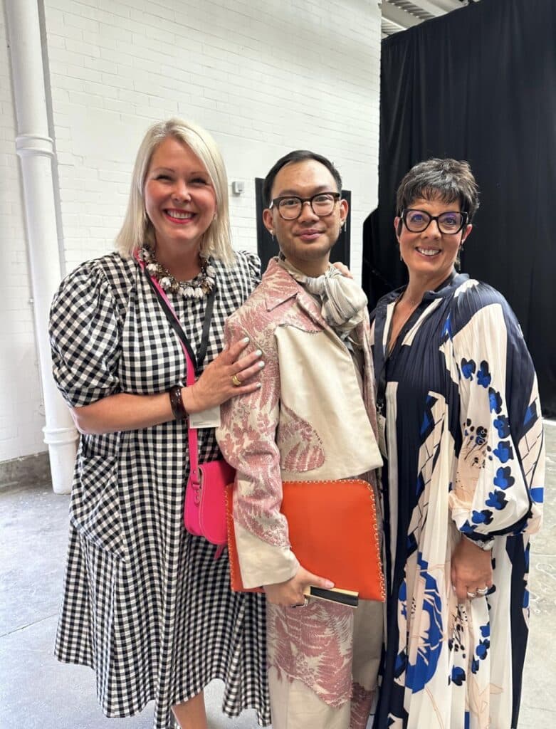 Three people stand together indoors, smiling. One wears a chequered dress, another a patterned suit, and the third a floral dress.