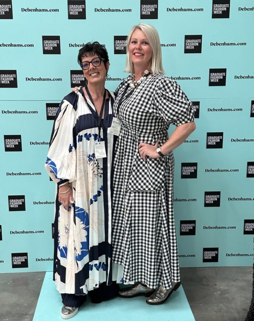 Two women stand smiling in front of a blue backdrop with "Debenhams.com" and "Graduate Fashion Week" logos. One wears a patterned dress with large flowers, and the other wears a black-and-white chequered dress.