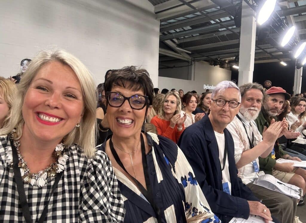 A group of people, seated closely together, smile and applaud at Graduate Fashion Week 2024. Some are wearing ID badges around their necks. The background shows more attendees and a section of the venue with bright lights.