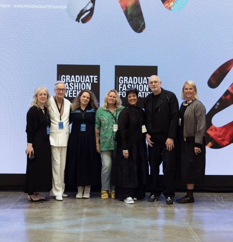 Group of seven people standing in front of "Graduate Fashion Week" banners, posing for a photo. They are wearing badges and smiling at the camera.