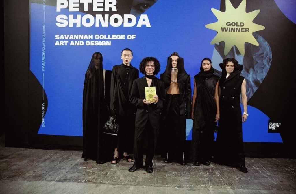 A group of six people in fashion outfits stand in front of a blue backdrop with the text "Peter Shonoda Gold Winner, Savannah College of Art and Design". One holds a gold award plaque.