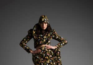 Person posing in a floral patterned outfit with a matching hood and mid-length skirt, standing against a plain grey background.