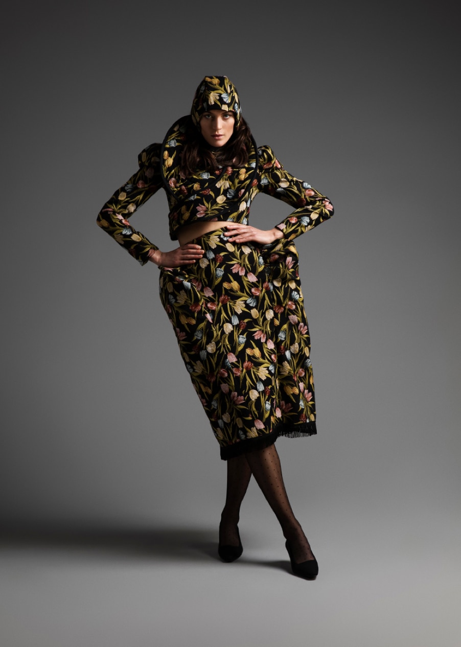 Person posing in a floral patterned outfit with a matching hood and mid-length skirt, standing against a plain grey background.