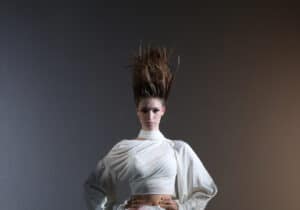 A person stands against a dark background wearing a light-coloured, avant-garde outfit with a high waist and dramatic headpiece. The lighting is dim, creating a moody atmosphere.