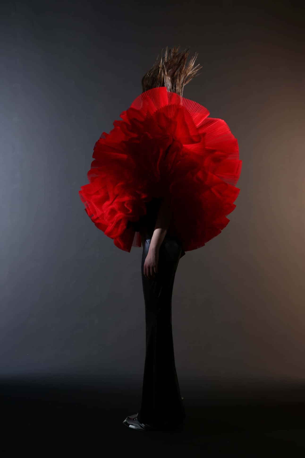 Person wearing a large, red, ruffled garment on the upper body and a long, black skirt stands against a dark gradient background. Hair is blown upwards.