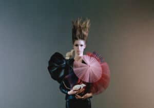 A woman with tall, upward-styled hair is wearing black attire with exaggerated shoulder pieces and holding red and pink handheld fans against a dark gradient background.