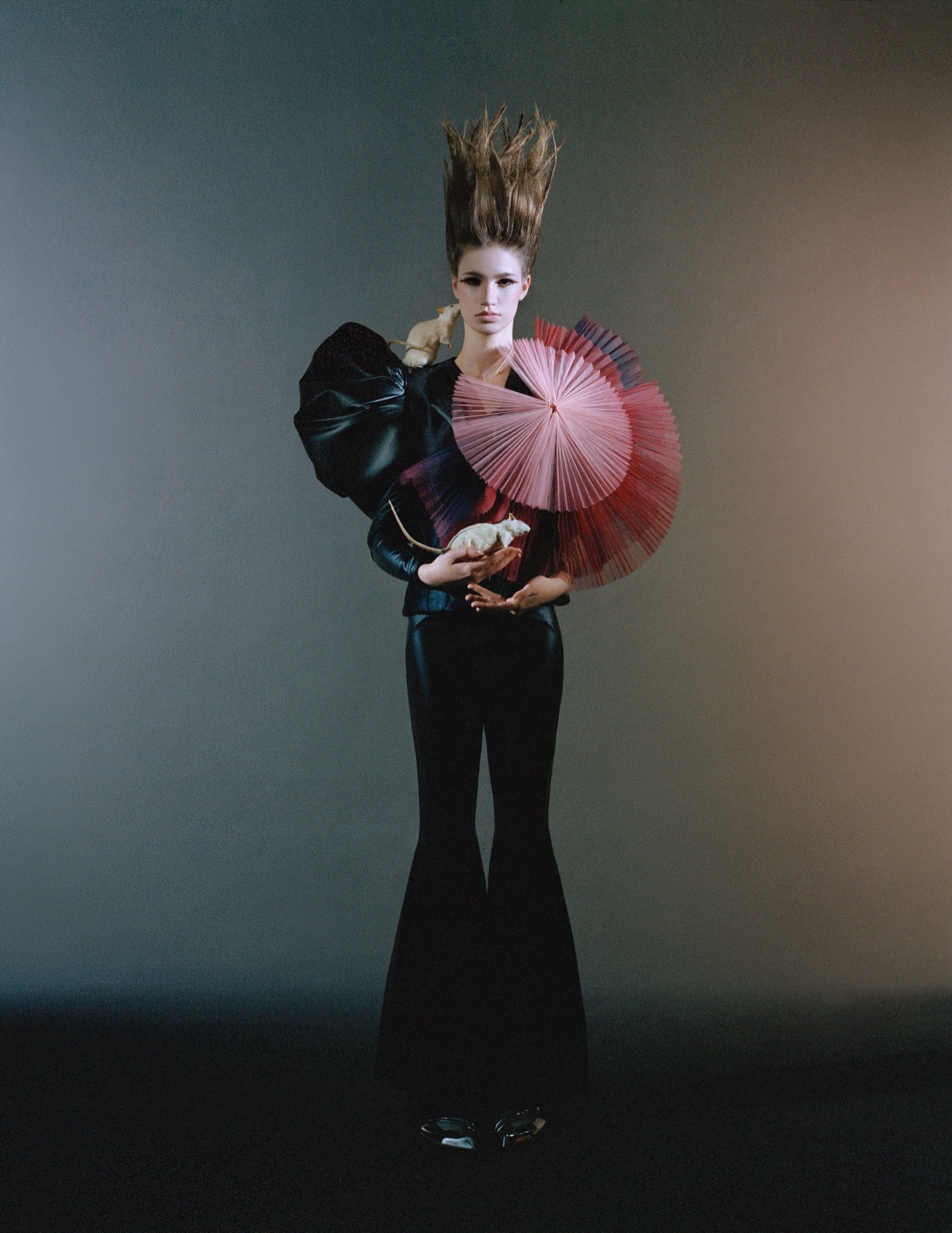 A woman with tall, upward-styled hair is wearing black attire with exaggerated shoulder pieces and holding red and pink handheld fans against a dark gradient background.