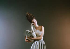 A model with windswept hair wears a strapless white dress embellished with lace, holding a large white swan against a dark gradient background.