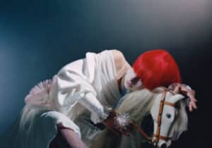 Person with bright red hair, white garment, and patterned tights posed on a wooden toy horse, under moody lighting.