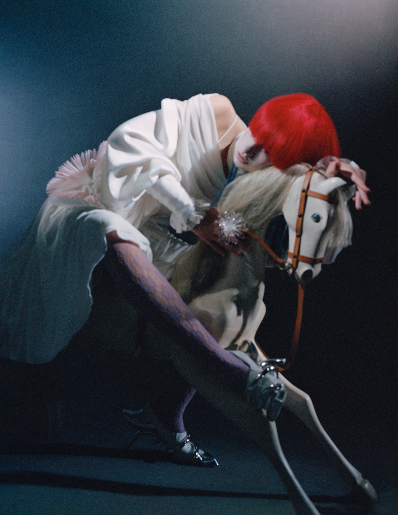 Person with bright red hair, white garment, and patterned tights posed on a wooden toy horse, under moody lighting.