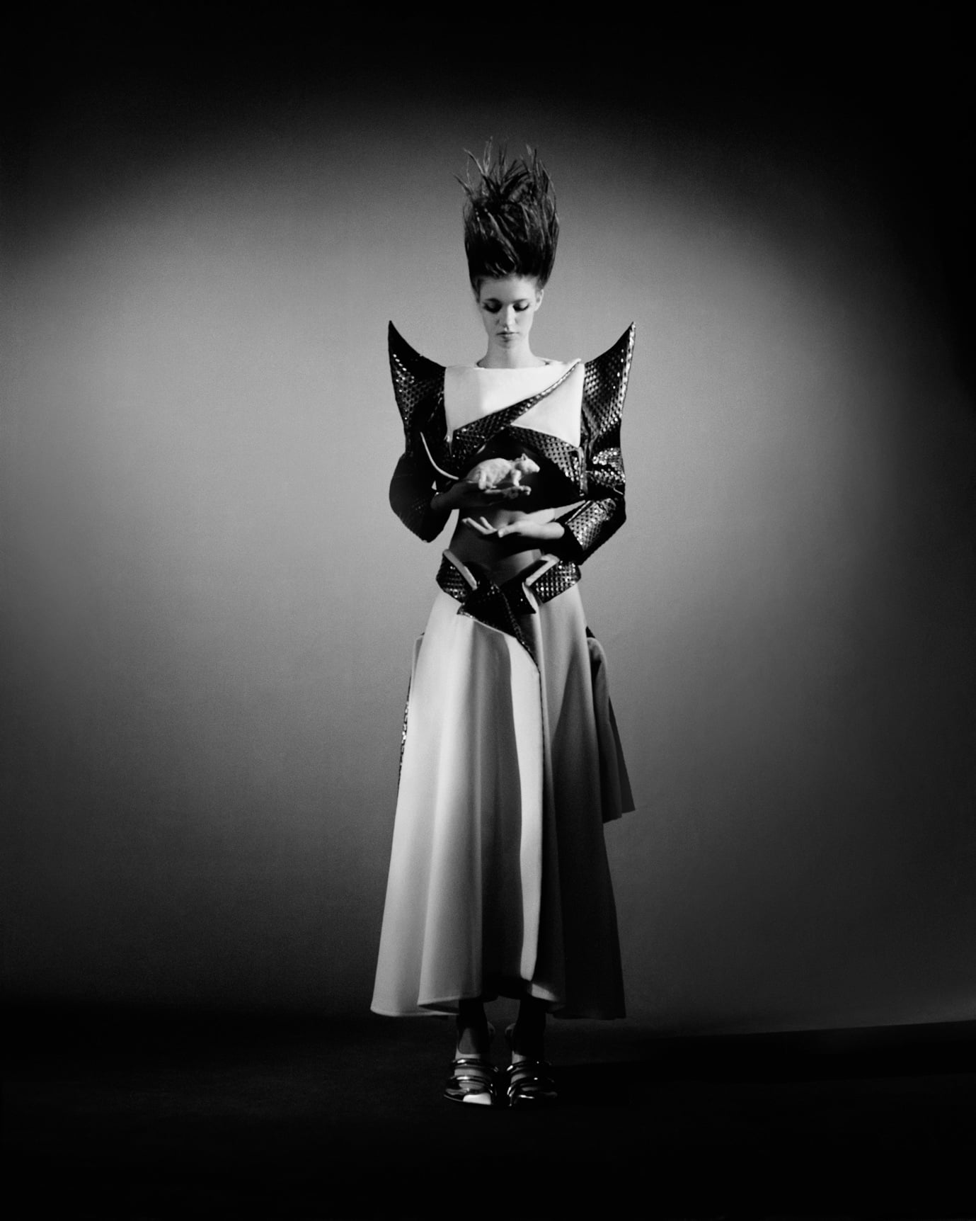 A woman stands in a futuristic dress with exaggerated shoulder features, holding a white rabbit against a dark background.